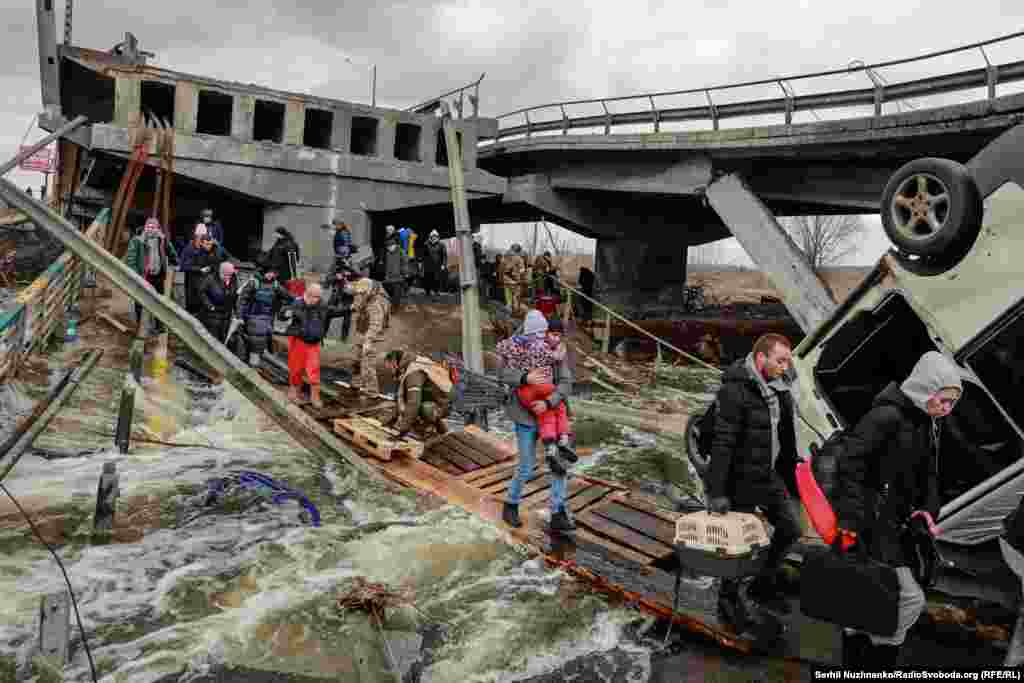 Людзі, несучы на руках дзяцей, пераходзяць раку па драўлянай кладцы пад падарваным мостам на мяжы паміж Ірпенем і жылым масівам Раманаўка. Сакавік 2022 году. Да 2022 году, паводле зьвестак Управы вярхоўнага камісара Арганізацыі абʼяднаных нацыяў па правах чалавека і ўкраінскіх праваахоўнікаў, у выніку баявых дзеяньняў загінулі 152 дзіцяці, 146 былі параненыя