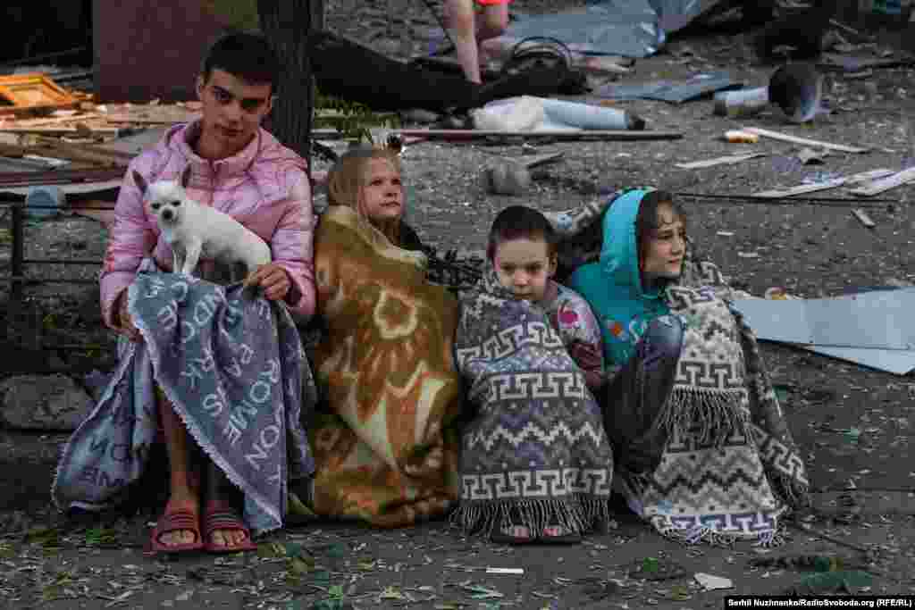 Дзеці, захутаныя ў коўдры, сядзяць разам з сваім сабачкам каля свайго дому, пашкоджанага ў выніку расейскай атакі. Жнівень 2025 году